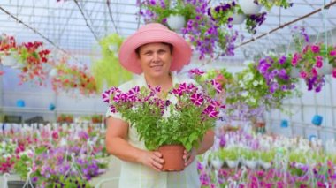 Front view nice senior female florist looking at camera stand in greenhouse smile hold hands pot of flower. Aged woman is happy, flowers in pots are growing beautiful, healthy and ready to sale.