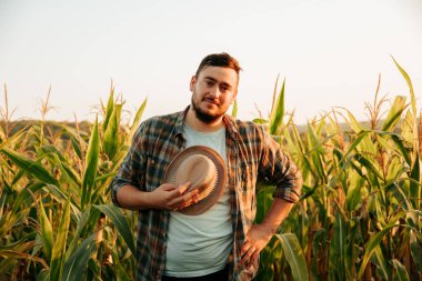 Pensive farmer with a smile on face stand in field, took off his hat, tired, front view, looking at camera. A young man on the background of a cornfield, one hand on hips, clear sky. Copy space.