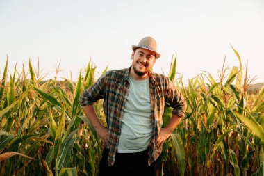 young man farmer stands in the field, smiles, keeps his hands on his hips, front view, looking at camera. The man is happy and shares his emotions. A rich cornfield is his job. Copy space.