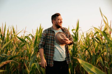 Background of a clear sky and a corn field, a young tired and contented male worker stands, took off hat and resting. It stands facing the sun, hold hat in hand. Front view, looking away. Copy space.