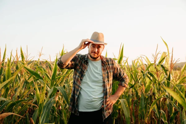 Young male farmer stands in field with one hand on hat, other hand on hips, front view looking at camera. The man greets everyone, smiling, background of a corn field and a clear sky. Copy space.
