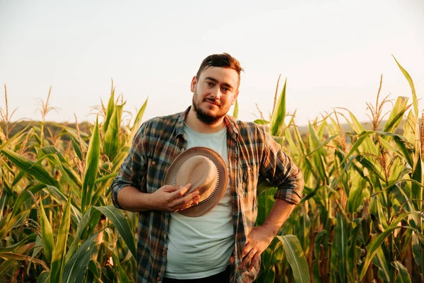 Pensive farmer with a smile on face stand in field, took off his hat, tired, front view, looking at camera. A young man on the background of a cornfield, one hand on hips, clear sky. Copy space.