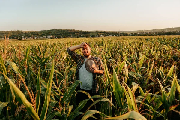 Tired young man stand in corn field, took off hat, wipes sweat from forehead, front view, looking away. Joyful farmer finished harvesting work, field background. Copy space.