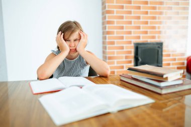 Front view looking at camera lazy student grimaces sitting in front of textbooks holding hands on head. There is a notebook and books on the table. She is in a room at her workplace. Copy space.
