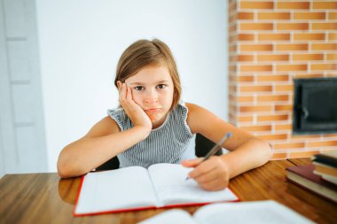 Front view looking at camera in room at desktop sits child girl grimace with displeasure hold head with hand. Elementary school student does not want to do homework. It is hard and bored. Copy space.