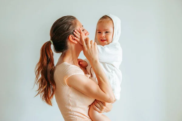 Background white wall young mom stand with child in arms baby hand on mother cheek on top woman arm. Kid dressed in a white bathrobe smiles happily. Side view infant mother. Copy space. Hand in hand.