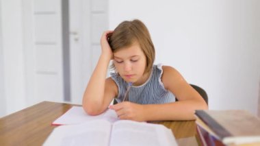 Front view schoolkid sits at desk, does homework with left hand looking at notebook. Student hold head with her hand, she is tired of doing homework. The child is bored. Copy space.