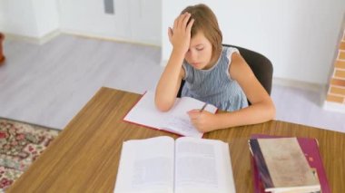 Top view schoolkid sit at desk write in copybook hold head with hand sigh heavily look in notebook. The student does her homework, she is hard and sad there are textbooks on the table. Child is bored.