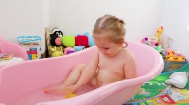 Front view baby girl sits in a baby pink bathtub plays with water, pouring it over a cup, smiles, turns her head. Little child is happy water calm down. Looking away, looking at camera.