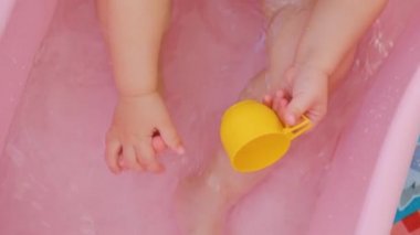 Close up top view unrecognizable feet and hands of a baby in a bath, plays with toes, a toy mug. The child sits in a bath of water, which splashes from the movements of the body.