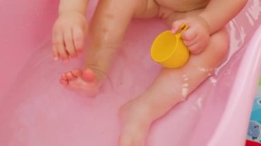 Close up unrecognizable baby feet hand in a splashing water plays with a small dinosaur and toes. The water in the bath splashes and pleases the child. In hand is a toy yellow cup bath of pink color.