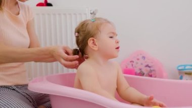 Front view playing in water mother, daughter in baby bath mom wash massaging child, girl smiles contentedly and turns her head. Children emotions of joy and happiness.