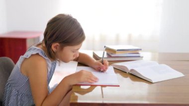 Side view school child girl student carefully, concentrate writes in notebook while sitting at table. Child is left hander, short hair, there are textbooks on table. Hard work doesnt let relax.