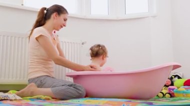  mom bathes her little daughter washes her back baby plays with water and turns her cup to face mother. Mum is happy talking. They are in large bright bathroom with white walls and lot of toys.