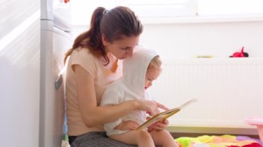 Little girl sits in mother arms listen attentively to an entertaining story. Mom points interesting things in book with finger. The baby child bounces a little in anticipation to go to play.