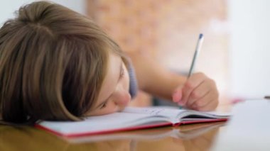 Front view close up child school girl head holding on the table writes lessons in a notebook with a pen. The student is doing homework at home. She is tired of doing homework, bored and lazy.