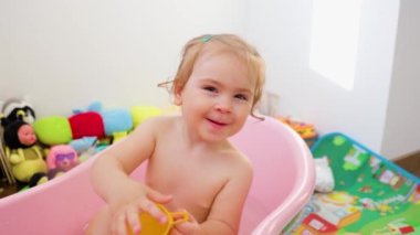 Front view little kid girl in a baby bath bathes plays with water laughs he stretches his hand towards the camera and looking at the camera. The child is happy, Carefree childhood and games.