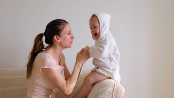 サイドビューお母さんの小さな赤ちゃんの娘は 母親が彼女の背中を持ち上げるダウンニット毛布の子供のスライドで覆われたソファでダンスを再生します 白いバスローブのママのカジュアルな服を着た女の子 白い壁だ スペースのコピー — ストック動画