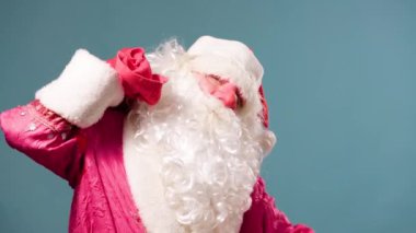Mavi arka planda Noel Baba 'nın kırmızı burnu hediyelerle dolu kırmızı bir çantanın altında duruyor. Yükünü kırmızı eldivenli elleriyle tutuyor. Kameraya bak..