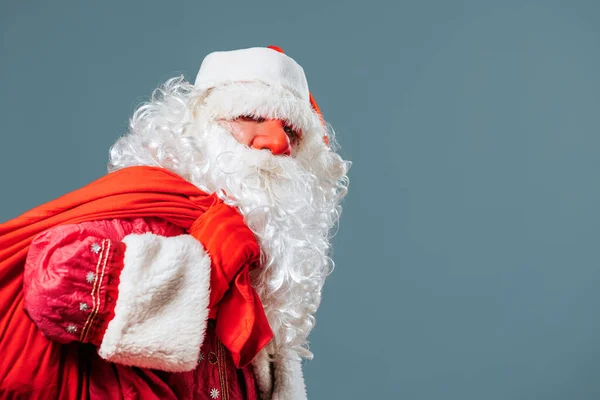 Santa Claus red nose on the blue background front view is leaning under the weight of a red bag with gifts. He holds his burden with his red gloved hands. Looks into the camera.