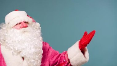 Mavi arka plan, Noel Baba şenlikli kameraya bakar, sonra yana doğru selamlaşırken elini sallar ve diğer eliyle bir şey gösterir. Weihnachtsmann, Pai natal, kopyalama alanı.