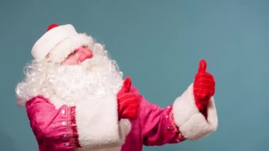 Şenlikli Noel Baba 'yı kaçıran mavi arka plan görüntüsü iki eliyle baş parmağını kaldırdığını gösteriyor. Kırmızı ve beyaz elbiseli mavi arka planda Noel Baba havalı görünüyor. Boşluğu kopyala.
