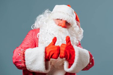 Noel Baba Pai natal mavi arka planda duruyor kameranın baş parmağına bakıyor ve bayramın yaklaşmasına seviniyor. Stüdyoda, Noel Baba seviniyor..