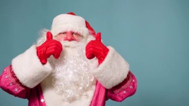 Noel Baba Pai natal mavi arka planda duruyor kameranın baş parmağına bakıyor ve bayramın yaklaşmasına seviniyor. Stüdyoda, Noel Baba seviniyor..
