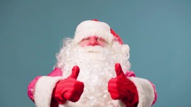 Noel Baba Pai natal mavi arka planda duruyor kameranın baş parmağına bakıyor ve bayramın yaklaşmasına seviniyor. Stüdyoda, Noel Baba seviniyor..