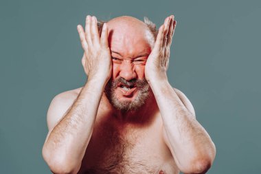 Kameraya bakan yaşlı bir adam, avuçlarını avuçlarının içine almış, farklı duygulardan yüzünü burkuyor ve sıkıyor. Stüdyo mavi arkaplan.