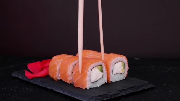 Chopsticks leva filadélfia rolo com peixe de salmão, abacate e queijo em uma placa preta no fundo escuro. — Vídeo de Stock