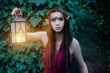 Portrait of a fabulous elven princess with a lantern in a green forest. Night in the forest. Magic and sorcery. Summer nature forest green tree. Long hair, pointed elf ears, green eyes, jeweled tiara.