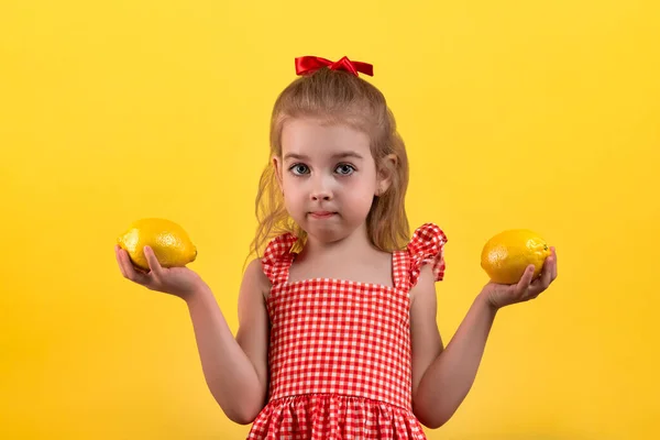 Cute girl with big eyes with lemons in her hands posing on a yellow background. Girl in a red plaid dress