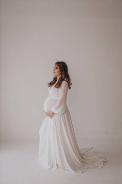 Genç, güzel, lüks, hamile bir kadın sağlam bir arka planda poz veriyor ve göbeğini gösteriyor. 