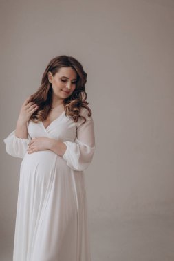 Genç, güzel, lüks, hamile bir kadın sağlam bir arka planda poz veriyor ve göbeğini gösteriyor. 