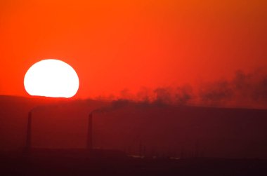The sun sets at sunset against the background of the pipes of the thermal power plant.