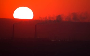 The sun sets at sunset against the background of the pipes of the thermal power plant.