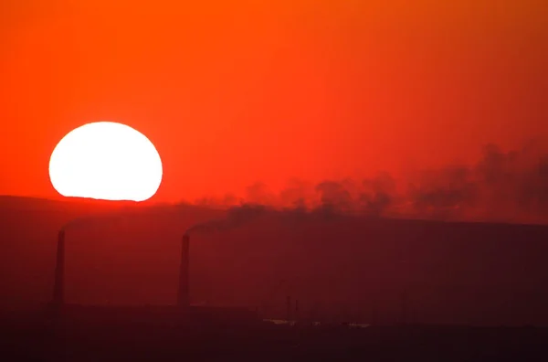 The sun sets at sunset against the background of the pipes of the thermal power plant.