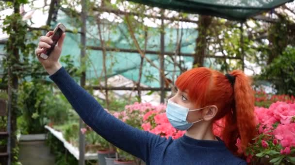 Ein Mädchen in einem Gewächshaus mit Maske fotografiert sich am Telefon vor dem Hintergrund von Blumen. Nahaufnahme — Stockvideo