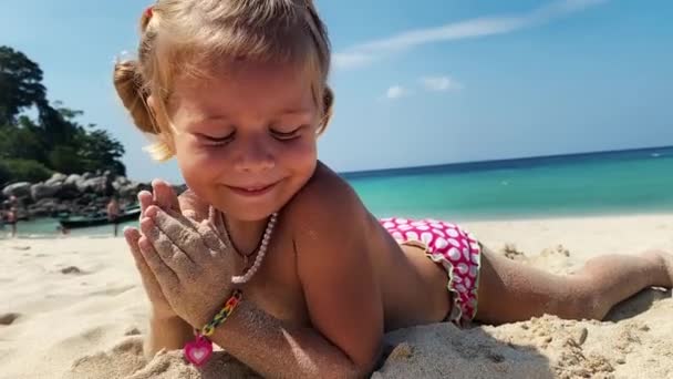 Klein meisje speelt met zand op het strand. Ze heeft paardenstaarten op haar, speelt met zand met handen en glimlacht. Kralen om haar nek, armbanden om haar handen. Vakantie met kinderen aan zee — Stockvideo