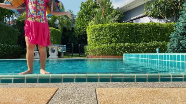 Bambina in costume da bagno rosa e maniche gonfiabili salta in piscina blu profondo. Schizzi dall'acqua si disperdono in direzioni diverse. I capelli biondi sono intrecciati in trecce. Buona infanzia. — Video Stock