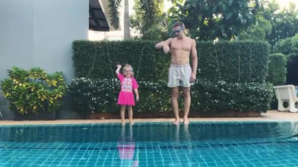 Vader leert dochter zwemmen in het zwembad. Papa en dochter steken hun handen op, papa springt in het water. Dochter in badpak en zwembril springt in zijn armen en zwemt. Gelukkige jeugd — Stockvideo