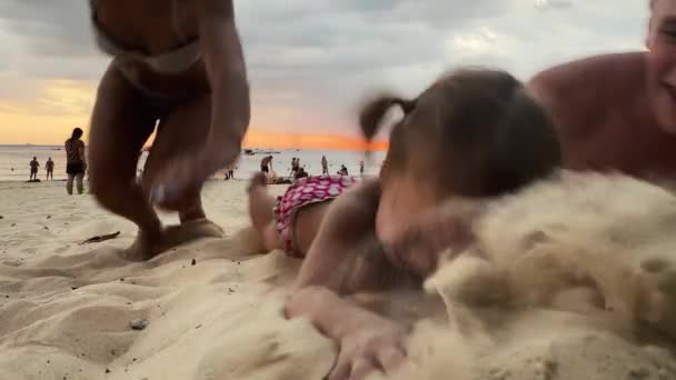 Mãe, pai e filha correm pela praia e caem na areia. A jovem família ri e sorri. Mamãe e papai beijam a filha na cabeça e ela ri. Feliz infância. Família. Amor. — Vídeo de Stock