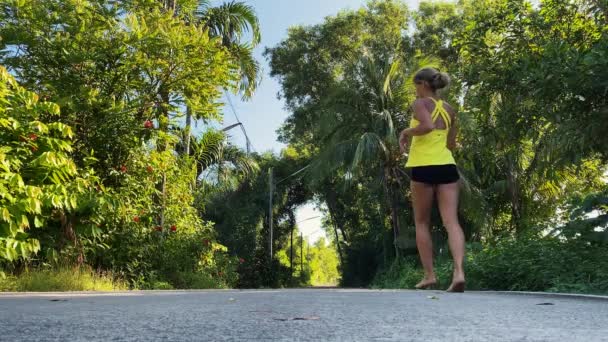 Genç bir kadın tropikal parkta fiziksel egzersiz yapıyor. Hamleler yapıyor. Sarı saçları at kuyruğu ile bağlanmış, sarı tişört ve siyah şort giyiyor, yalınayak. Eğlence için spor — Stok video