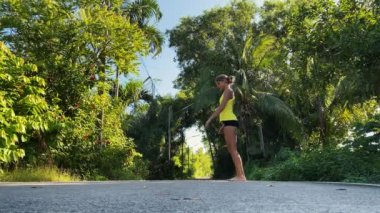Kadın tropikal parkta fiziksel egzersiz yapıyor. Hamleler yapıyor, geriniyor ve zevkle zıplıyor. Saçları at kuyruğuyla bağlanmış, sarı tişört ve siyah şort giyiyor. Eğlence için spor