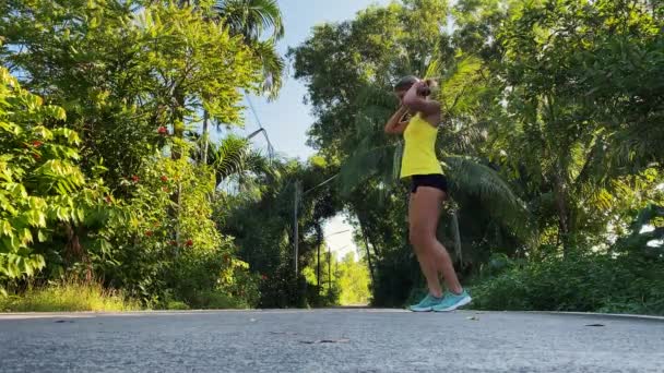 Kadın tropikal parkta fiziksel egzersiz yapıyor. Zevkle hamleler yapıyor. Saçları at kuyruğuyla bağlanmış, sarı tişört ve siyah şort giyiyor. Eğlence için spor — Stok video