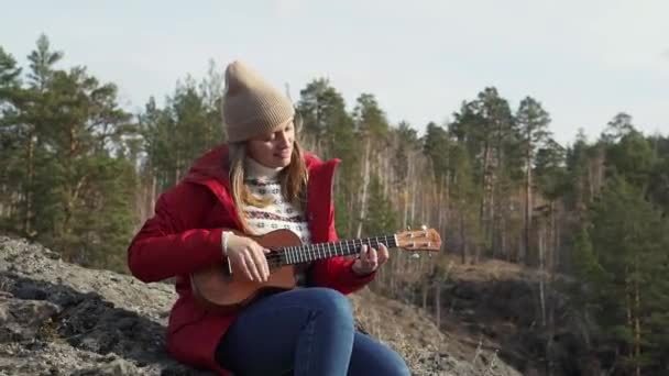 백인 여성은 바위 위에 앉아 우쿨렐레를 연주 한다. 그녀의 금발 머리는 바람에 날려 갑니다. 그녀는 작은 기타줄을 연주 한다. 가을, 나무 위의 노란 잎들. 취미, 현악기, 음악 — 비디오