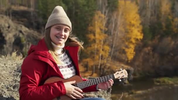 A mulher senta-se na rocha e segura o ukulele. Seu cabelo loiro é soprado pelo vento. O sol está brilhando e ela está sorrindo. Outono, folhas amarelas nas árvores. Passatempos — Vídeo de Stock