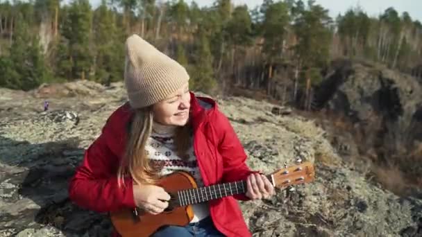 Kobieta siedzi na skale i gra na ukulele. Jej blond włosy wieje wiatr. Portretuje gwiazdę rocka i aktywnie uderza w struny i śpiewa. Jesień, żółte liście na drzewach. Zainteresowania — Wideo stockowe