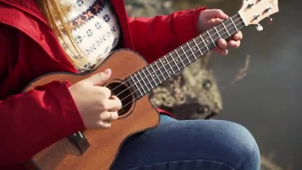 La donna caucasica siede sulla roccia e suona l'ukulele. I suoi capelli biondi sono soffiati dal vento. Suona le corde di una piccola chitarra. Autunno, foglie gialle sugli alberi. Interessi, stringa, musica — Video Stock